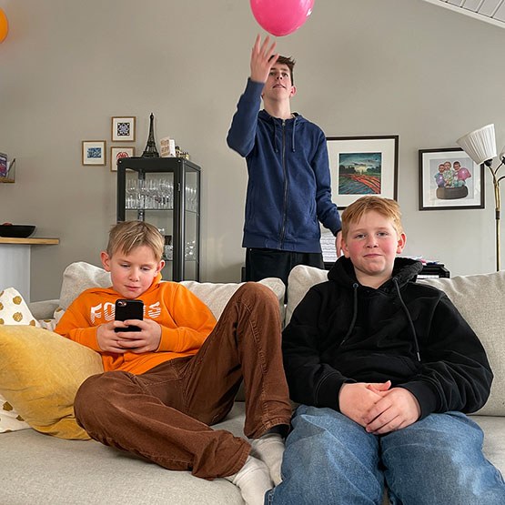 Den unge sitter i sofaen. Lillebror sitter ved siden av og trykker på mobil. Storebror står bak og leker med en ballong.
