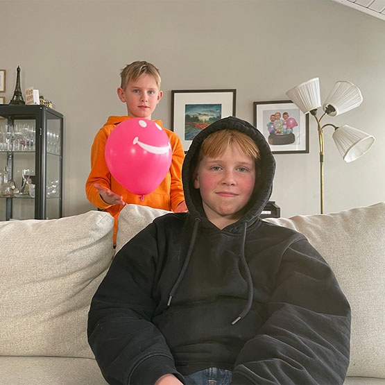 Den unge sitter i sofaen med hetta på. Lillebror står bak og leker med en ballong.