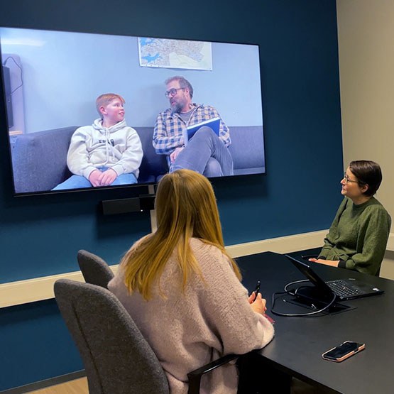 To voksne sitter på møterom og ser på skjermen. På skjermen er terapeuten og den unge.